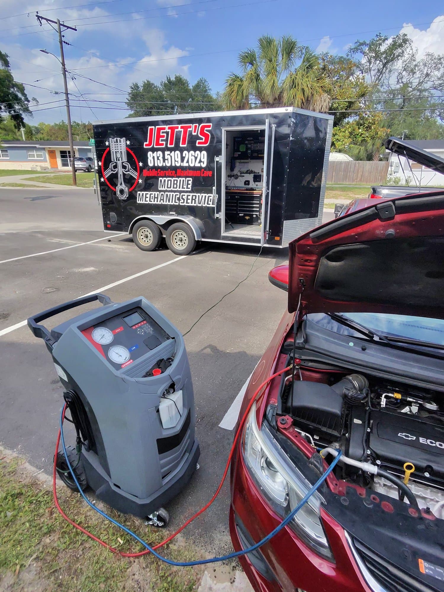 The A/C recovery machine in a parking lot with its red and blue hoses connected to a dark red Chevy with the hood up, the open trailer parked behind