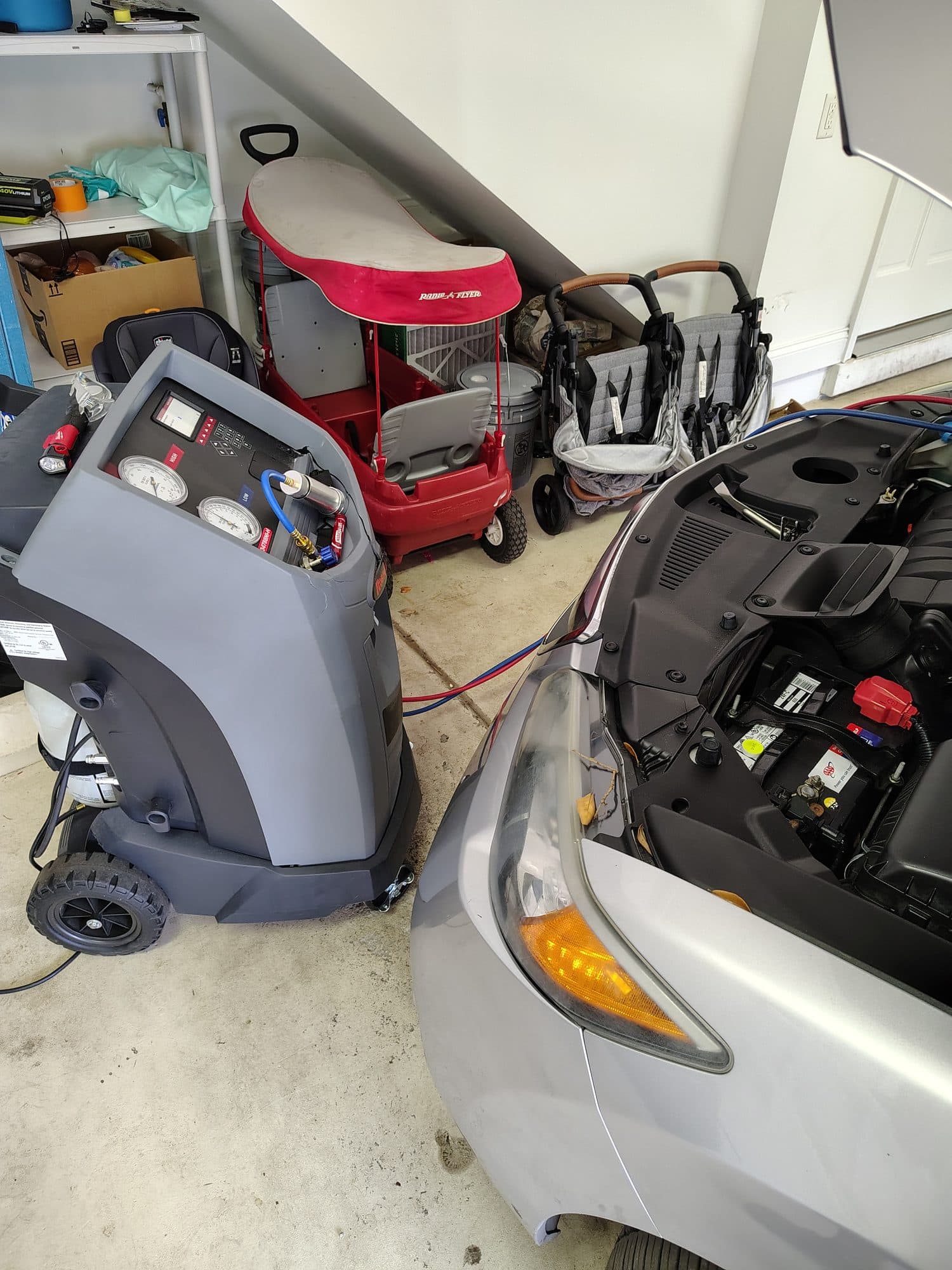 The A/C recovery machine wheeled into a garage with its red and blue hoses connected to a silver car's air conditioning ports, hood open