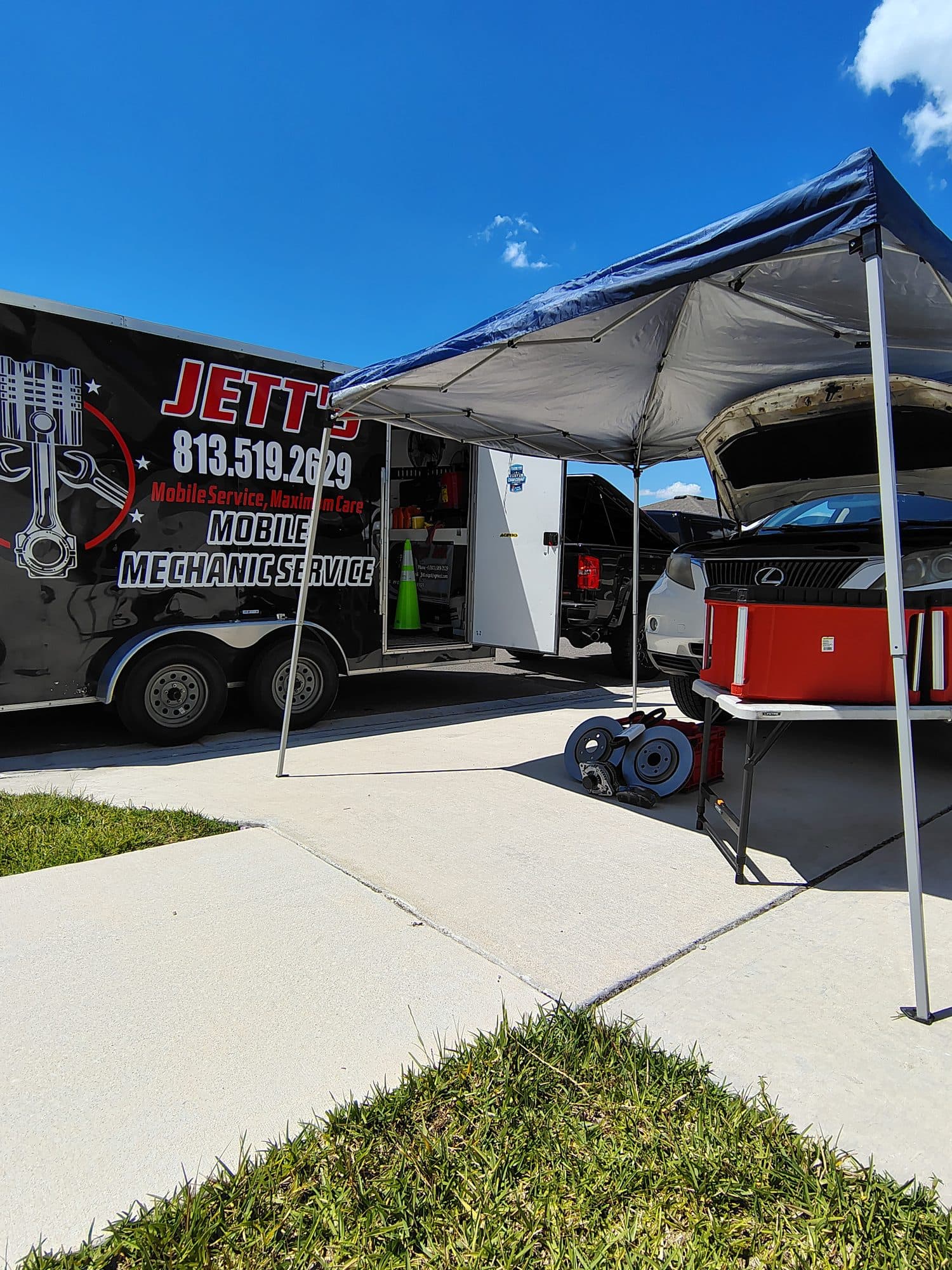 A pop-up canopy set up beside the open trailer on a driveway, shading a Lexus SUV with its hood up and a pair of brake rotors on the work table