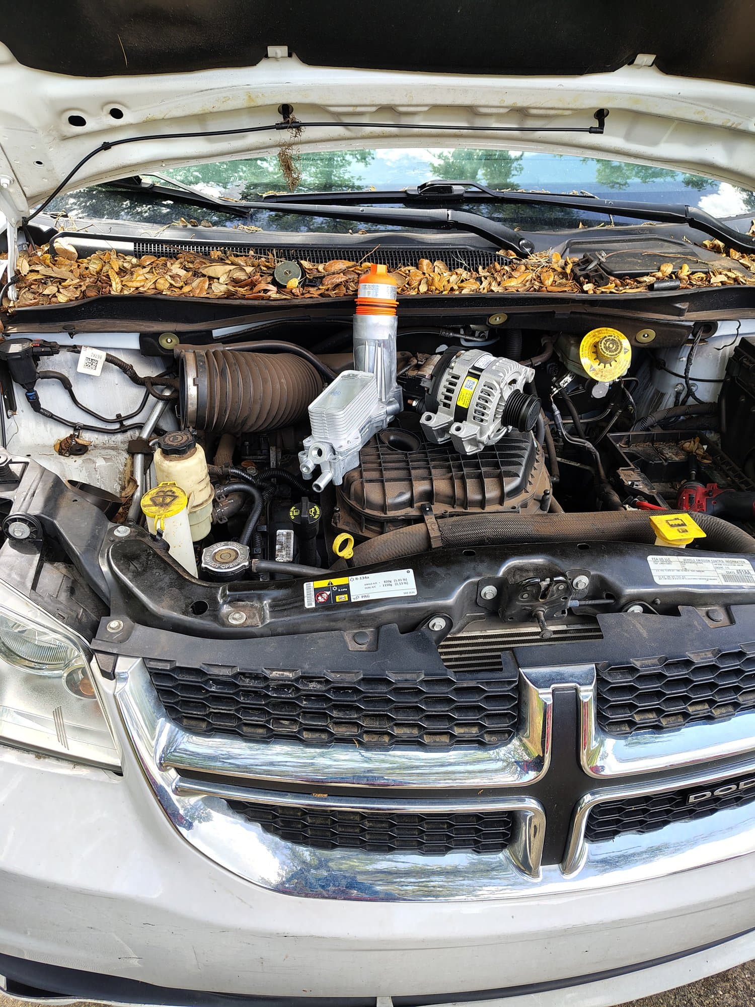 Under the hood of a white Dodge with the front end opened up, a brand-new alternator fitted at the top of the engine with its yellow sticker still on