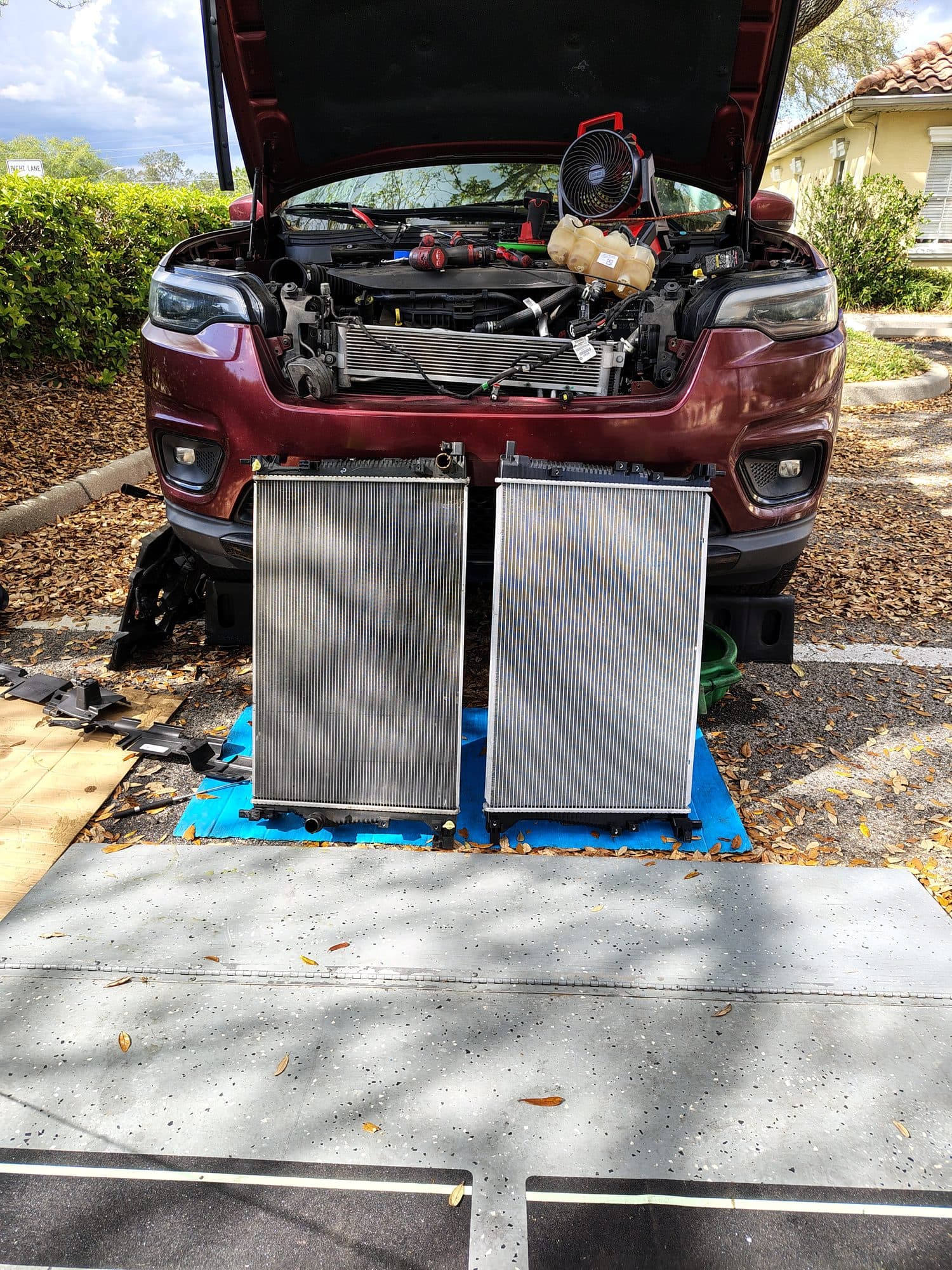 A dark red Jeep with the front bumper and grille removed, the old and new radiators standing side by side on the ground in front of it