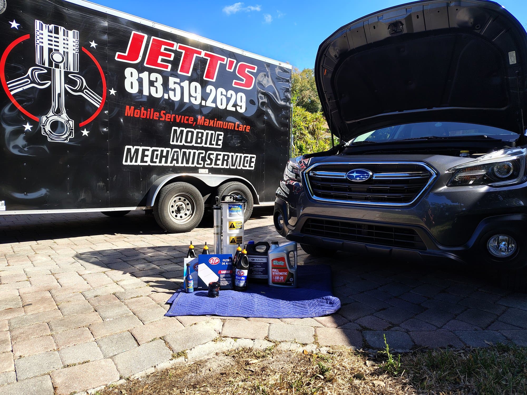The trailer parked beside a grey Subaru Outback with its hood up on a paver driveway, the fresh oil, filter and supplies laid out on a blue moving blanket in front of the bumper