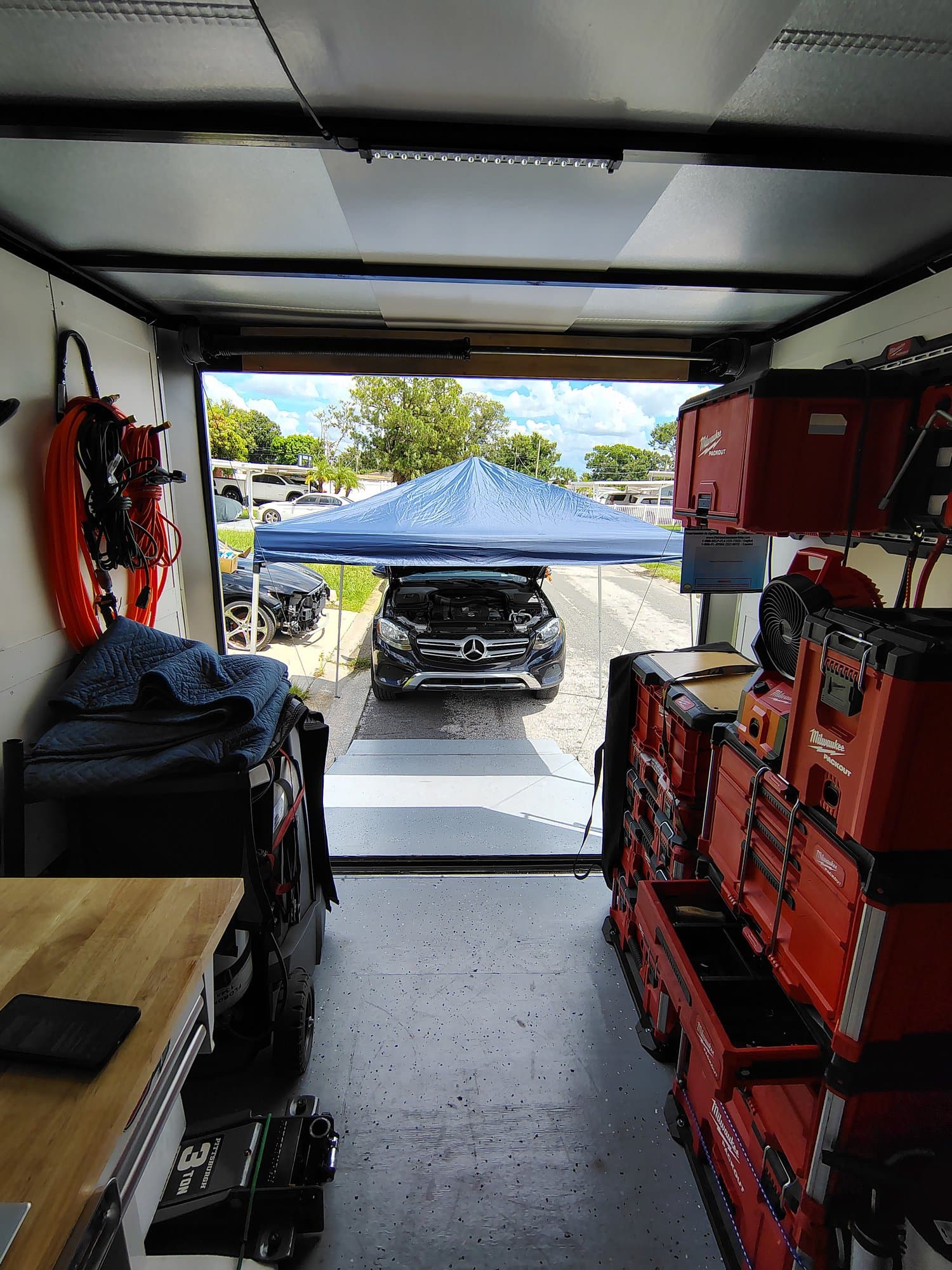 Looking out from inside the trailer, past the workbench and the wall of red tool cases, to a blue canopy shading a dark Mercedes SUV with its hood up in the driveway