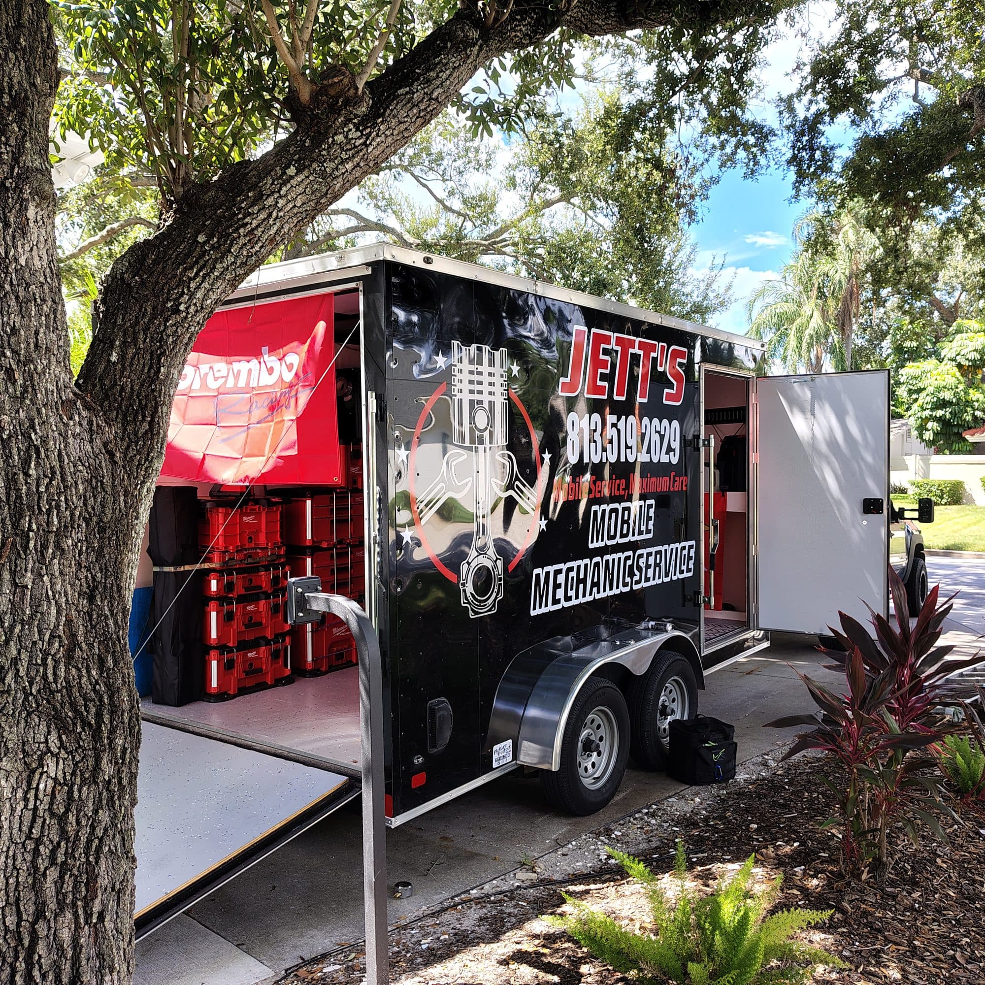 The trailer parked under a live oak with its ramp down and side door open, showing stacked red tool cases and a Brembo banner inside
