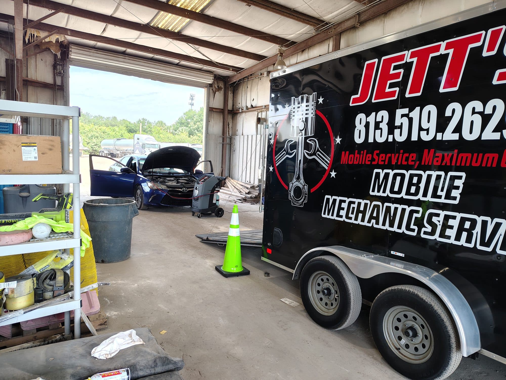 The trailer backed into an open shop bay, with a blue Toyota sedan on the far side, hood up and a service cart pulled alongside