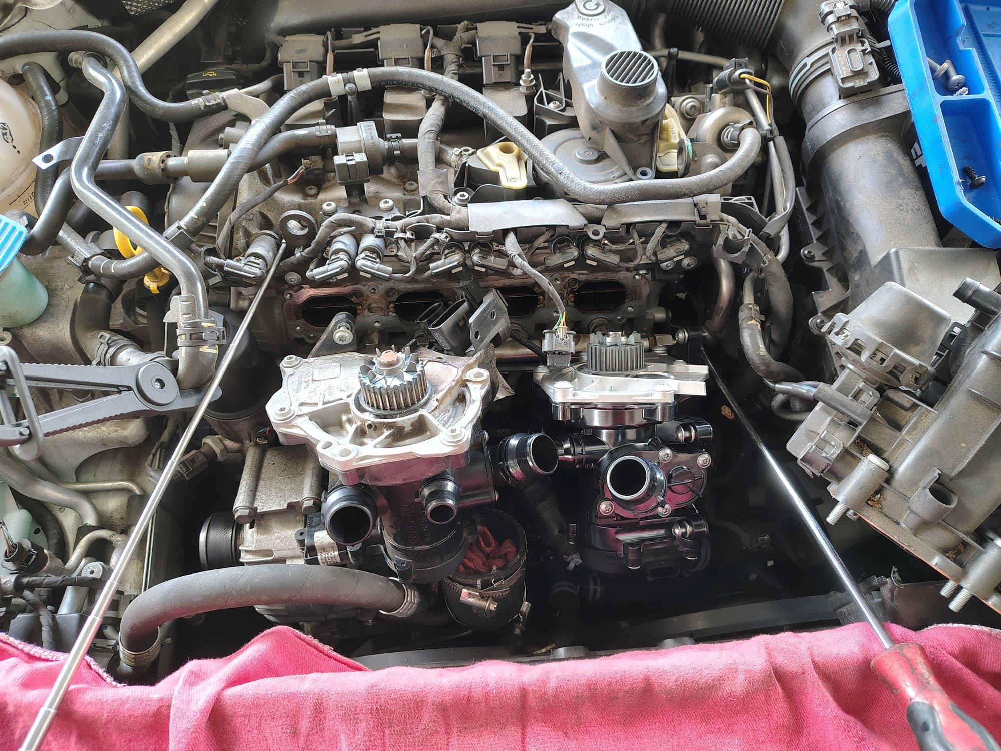A Volkswagen engine with the intake manifold removed to reach the water pump, the old pump and thermostat assembly and the new one sitting side by side on the block